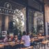 Atmospheric exterior view of a cozy cafe with wooden tables and a person working on a laptop.